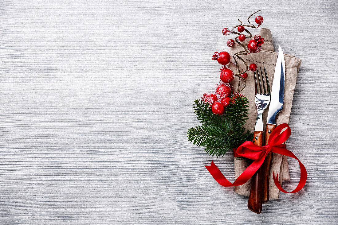 Table fork and knife set with napkin, Christmas fir branch, red berries and ribbon on grey wooden background Copy Space