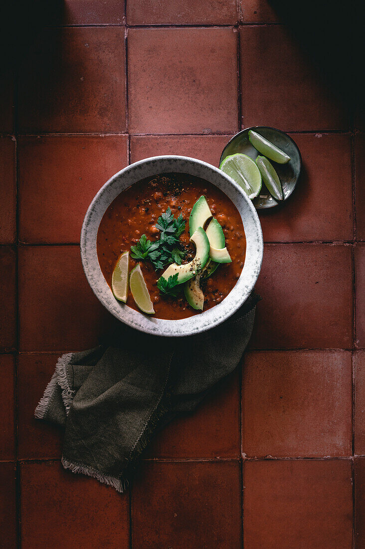 Chili protein bowl with avocado slices, fresh lime and parsley in a red terracotta surface in a moody style.