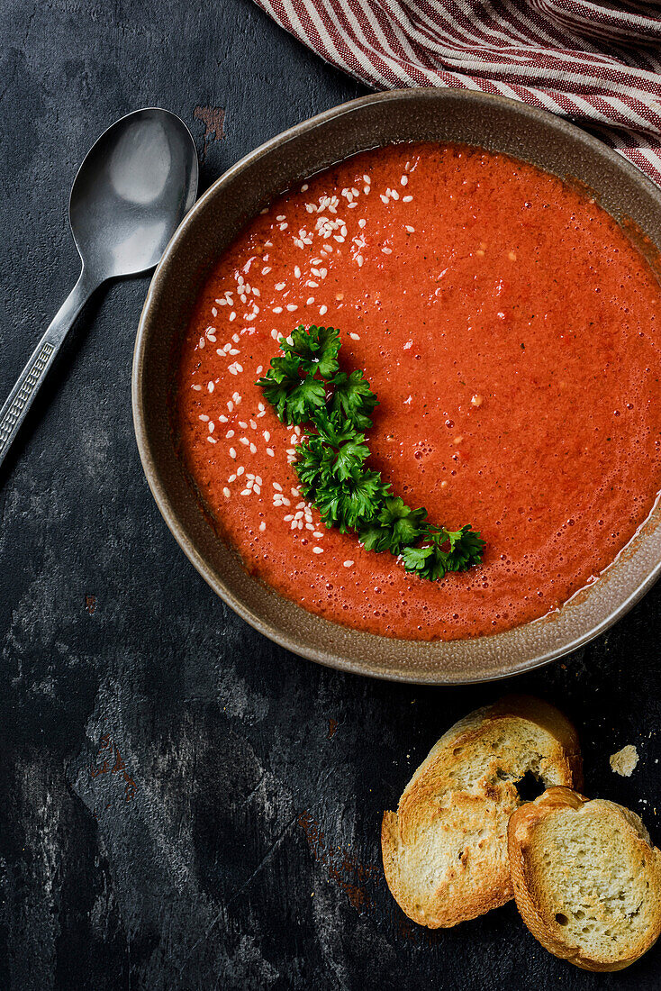 Gazpacho garnished with parsley in a plate with a wavy edge. On the table are red peppers, tomatoes, garlic, croutons and a spoon. View from above