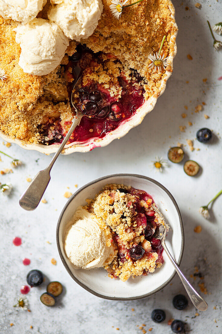A serving of apple and blueberry crumble in a bowl with a scoop of vanilla ice cream and the remainder of the pudding in the baking dish in view.