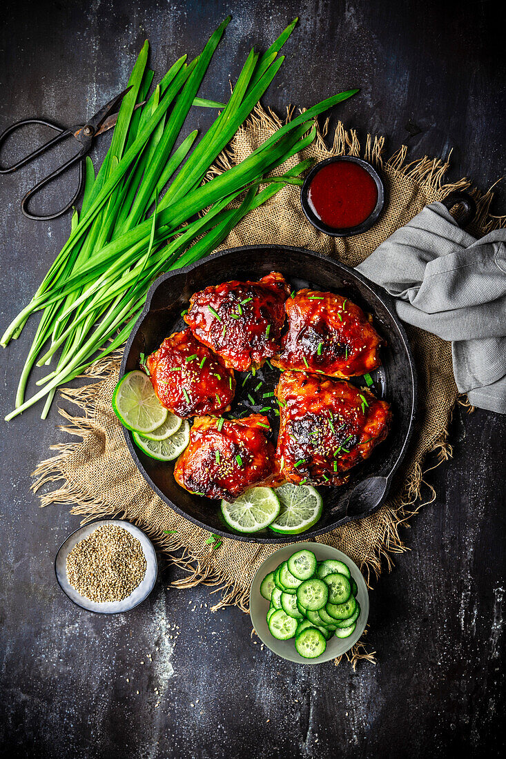 Vintage cast iron pan with spicy glazed chicken thighs with sauce, sliced cucumber and chives, overhead