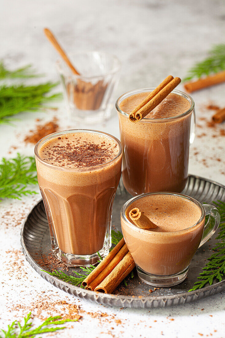 Three glass mugs in various sizes holding hot chocolate with cinnamon sticks as a garnish.