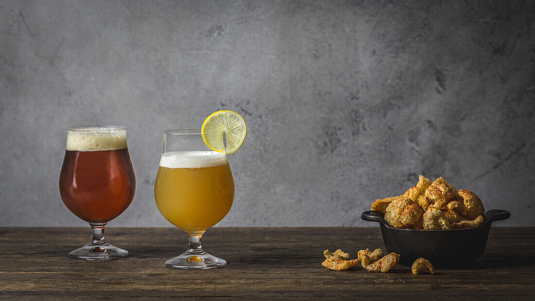 A wheat beer and a brown beer, in tulip glasses on a wooden table with a bowl of pork rinds