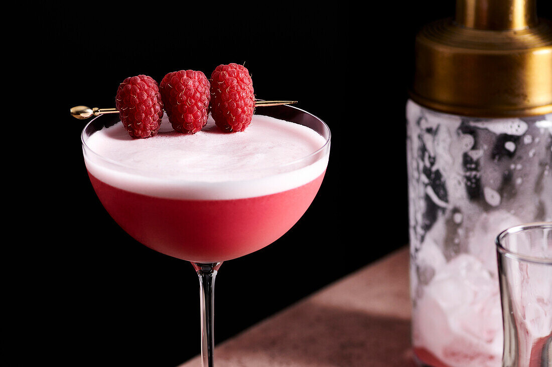 Clover club cocktail in a tall coupe glass, garnished with three raspberries on a golden pick