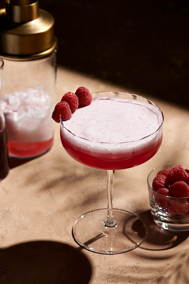 Clover Club cocktail in a tall coupe glass, garnished with three raspberries, next to a shaker in hard light