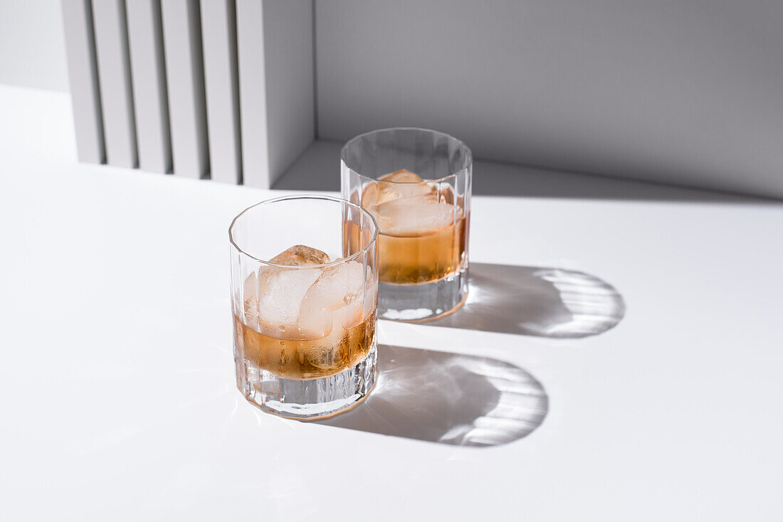 High angle of clear glasses filled with cold refreshing scotch whiskey with ice cubes on white surface
