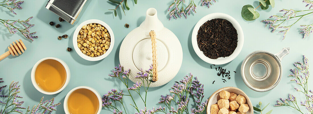 White porcelain teapot, two cups, sugar and a selection of dry tea in white ceramic bowls on a blue background. View from above. Banner