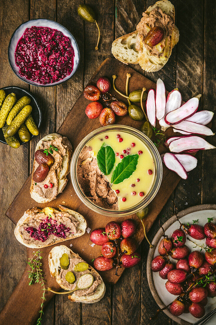 Pastetenaufstrich im Glas auf Holzbrett mit gerösteten Trauben, Radieschen, Essiggurken und Baguette