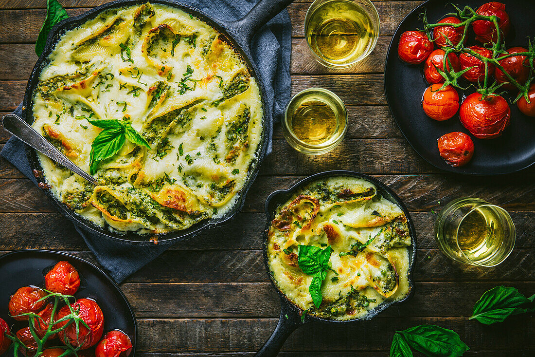 Small and large cast-iron pan with baked cheese spaetzle, garnished with roasted tomatoes, wine and basil on a wooden table