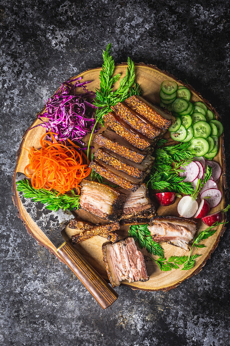 Sliced crispy pork belly on a wooden plate with a knife and colourful vegetable garnishes
