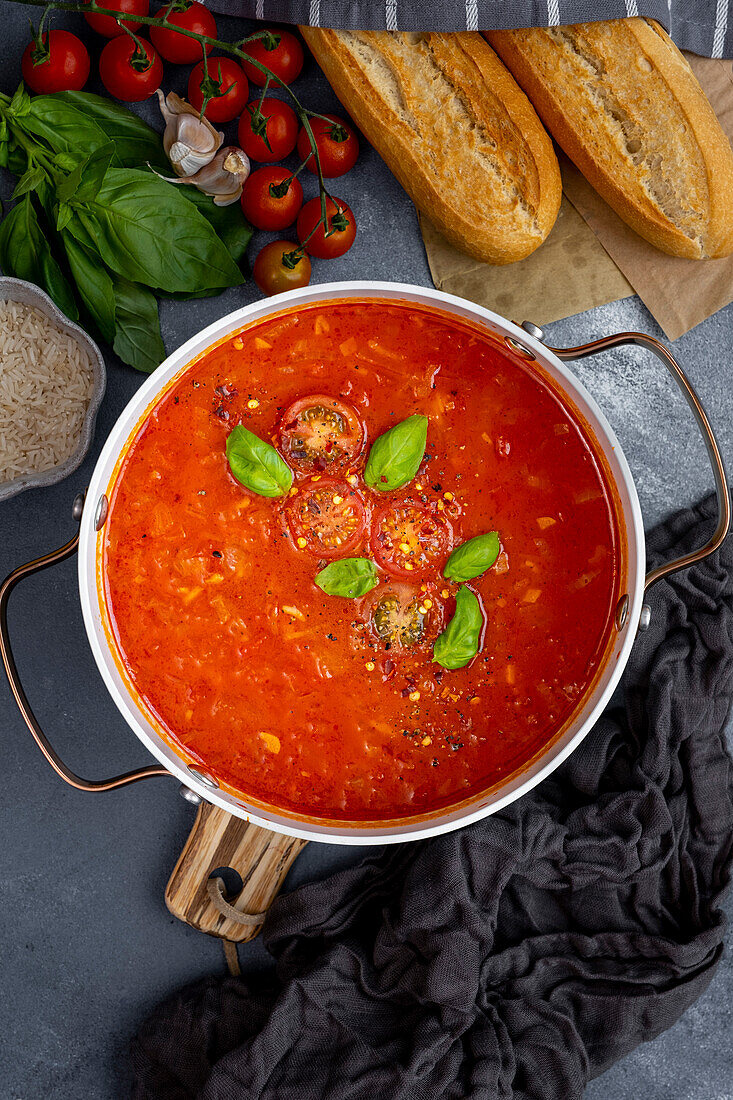 Tomato and rice soup garnished with halved cherry tomatoes and basil leaves in a white pot, two pieces of bread, cherry tomatoes, garlic and basil leaves on the side