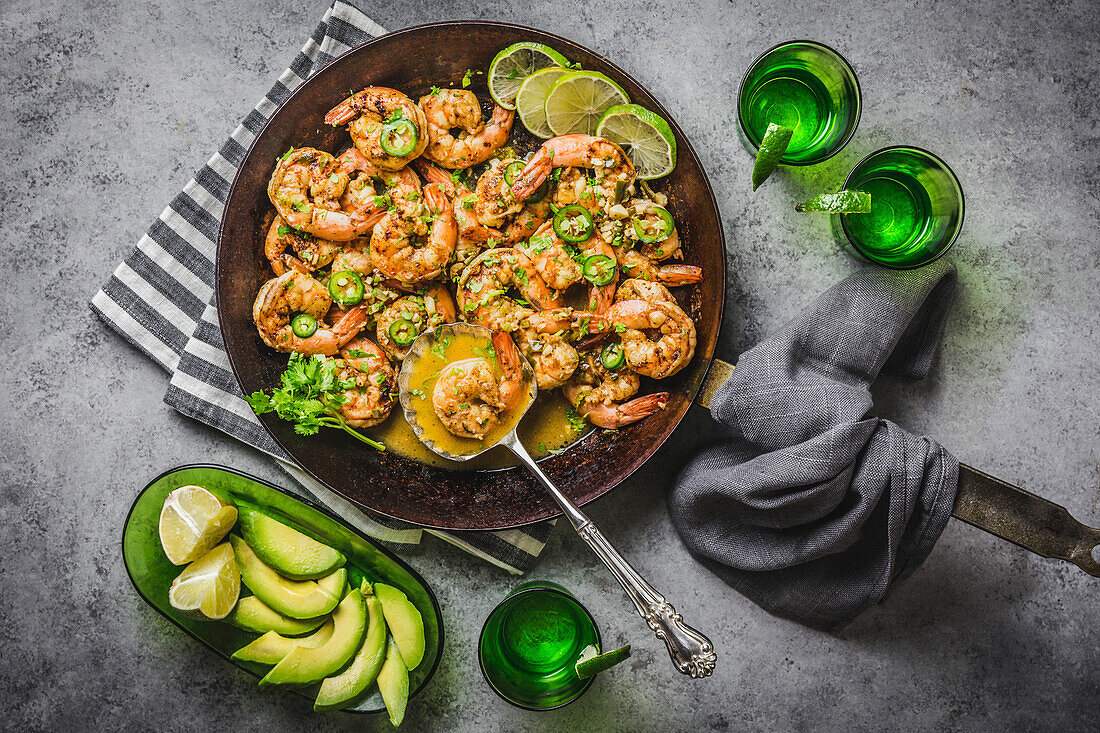 Prawns with chilli, lime and coriander in butter sauce, carbon steel pan, with avocado slices on grey linen napkins and silver spoon