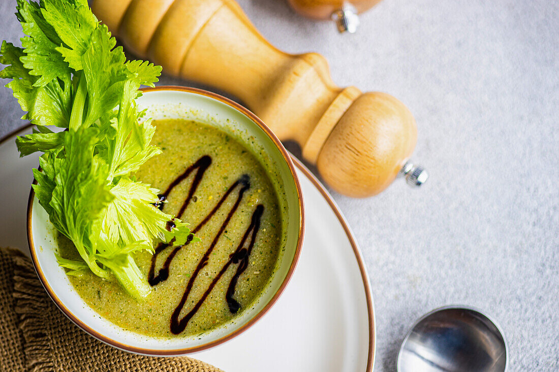 Top view of healthy celery cream soup in bowl with celery sticks on white plate with napkin on grey table with spoon and salt and pepper wooden jars