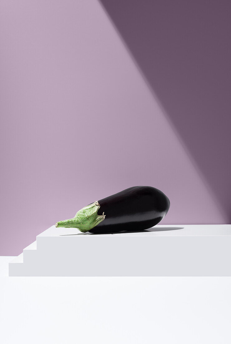 Vivid image of a black aubergine with a green cap lying on a white tiered plate against a purple background under bright light