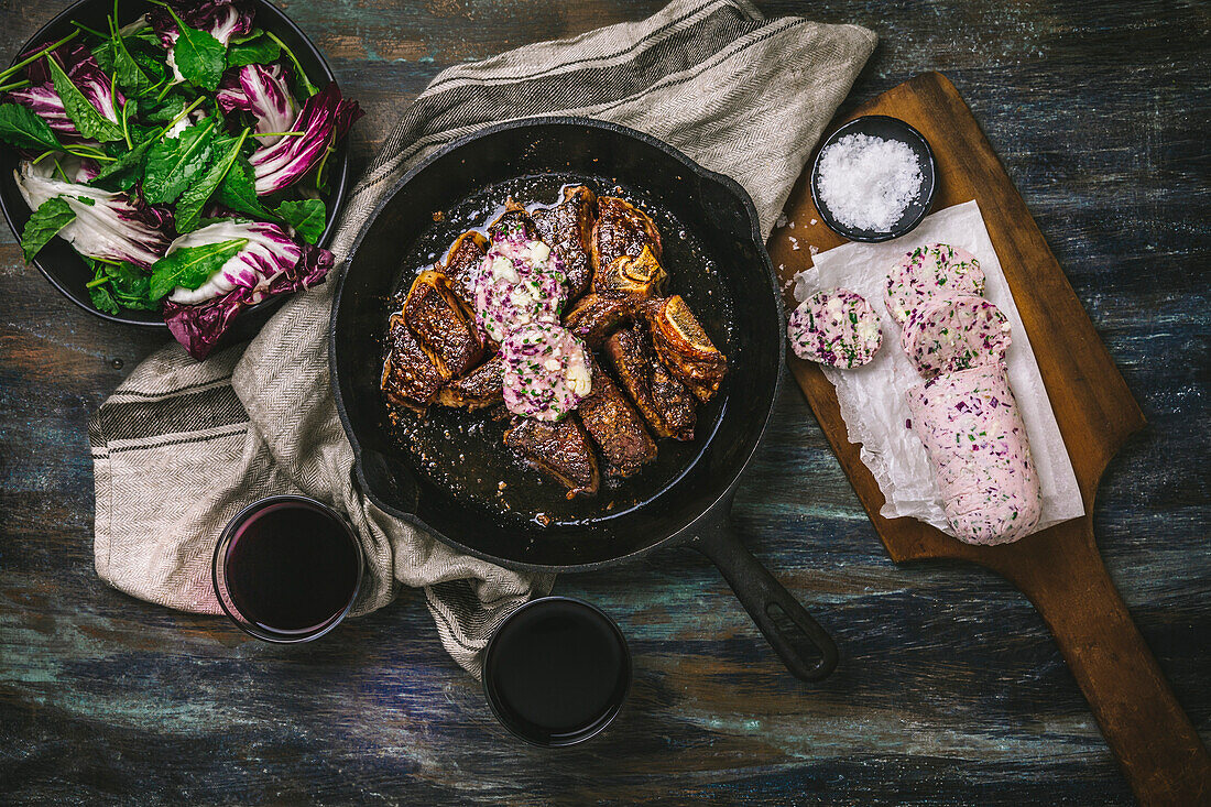 T-bone sirloin steak with red wine butter in a cast iron pan with butter slices on the board, salad and wine glasses