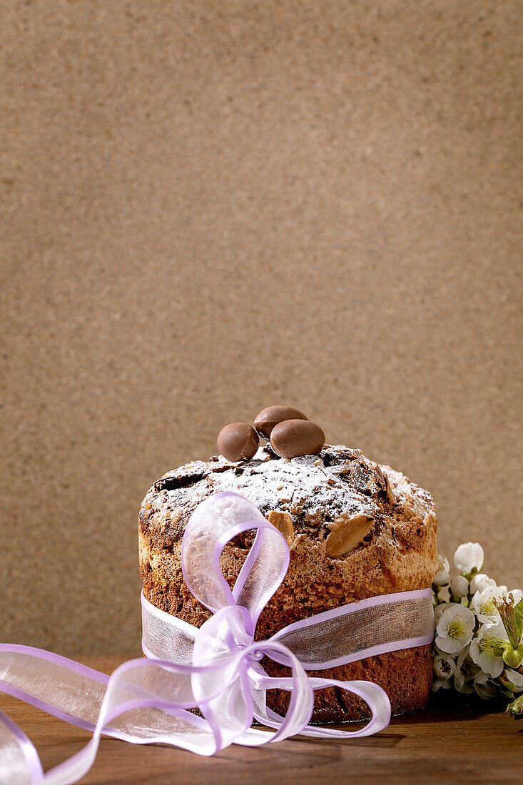 Hausgemachter italienischer traditioneller Oster-Panettone-Kuchen, verziert mit Schokoladeneiern, rosa Schleife und blühenden Kirschbaumblüten auf Holztisch stehend. Traditionelles europäisches Ostergebäck. Platz zum Kopieren