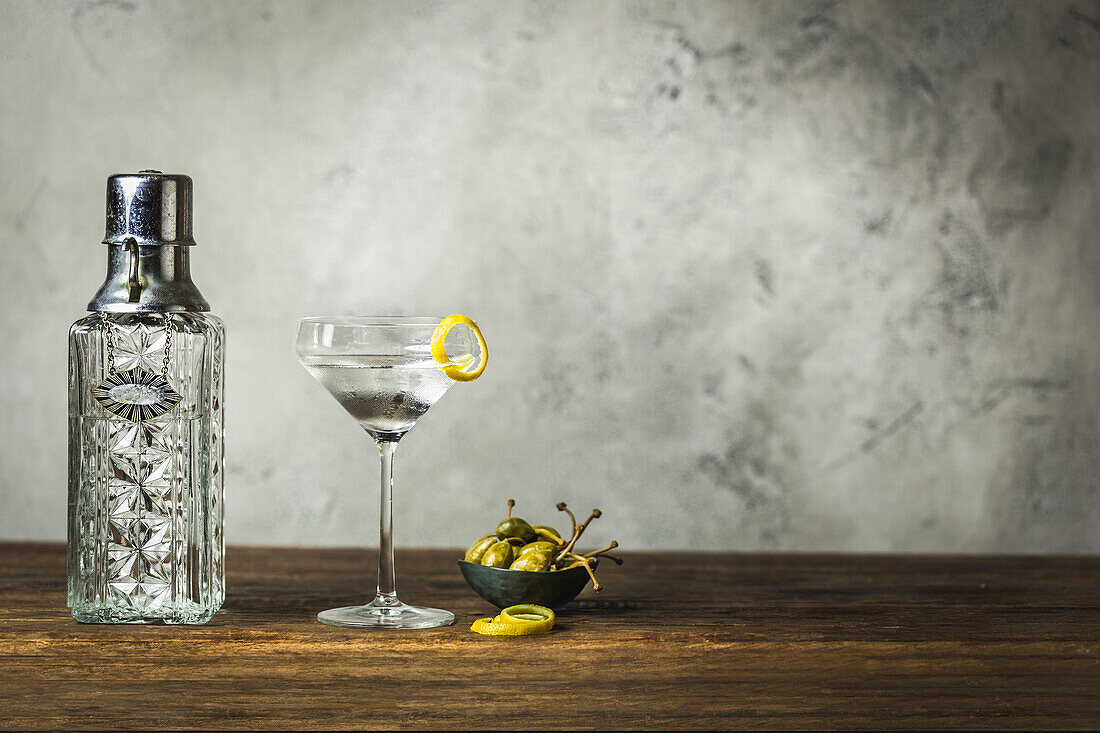 Martini with lemon twist, vintage decanter, caper berries in a small bowl