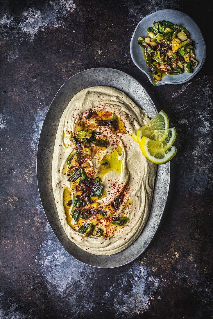 Hummus on an oval metal plate tossed with charred spring onions, lemon slices and olive oil
