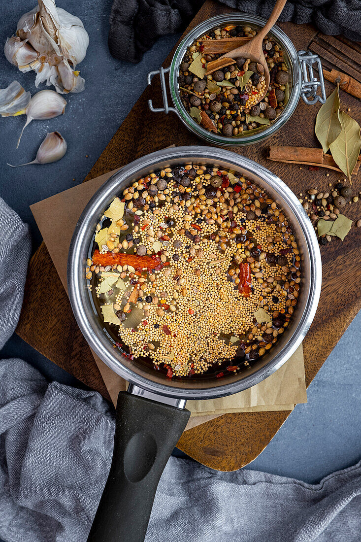 Pickling brine with mustard seeds, coriander seeds, cinnamon sticks, bay leaves, cloves, allspice berries in a saucepan on a wooden cutting board.
