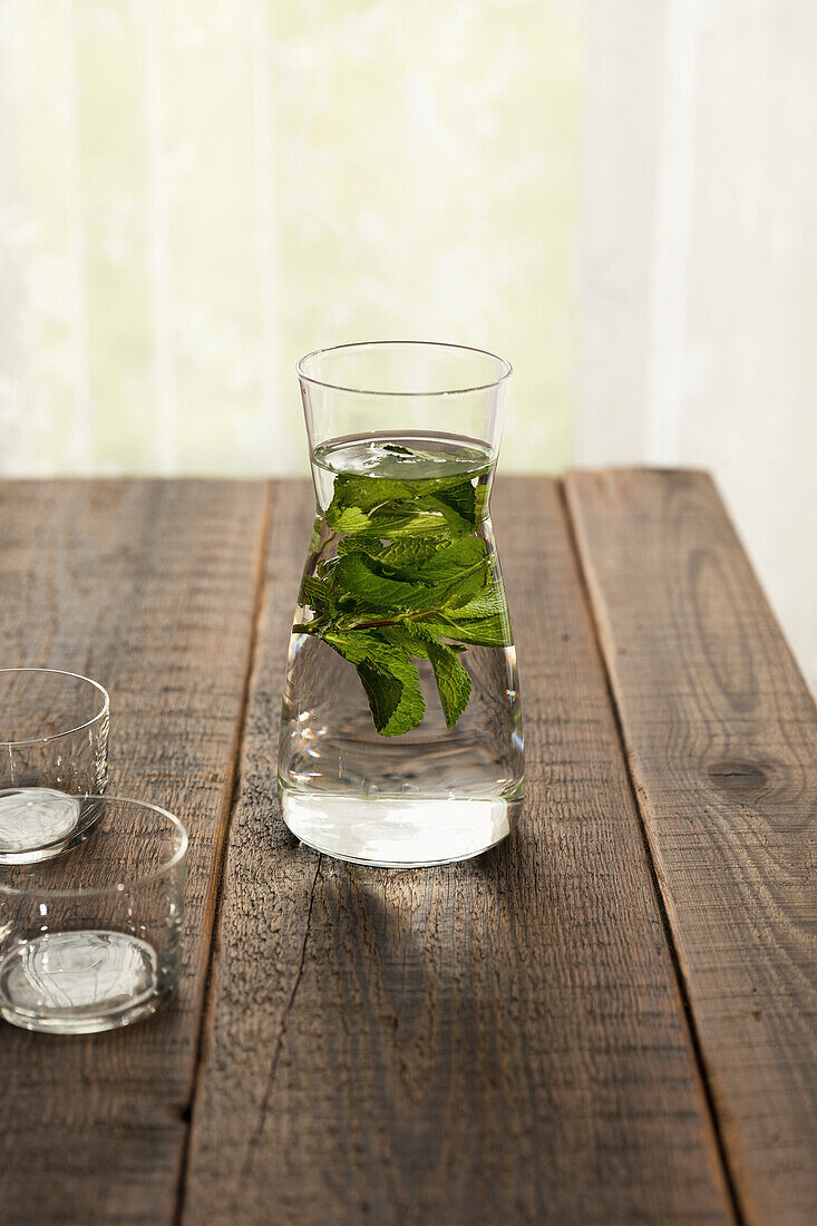 Fresh water and mint leaves in a carafe and two cups filled with water, on a wooden table