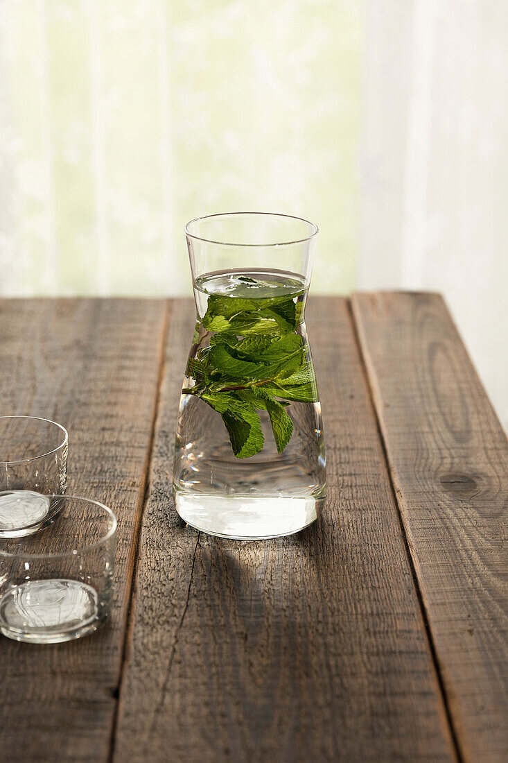 Frisches Wasser und Minzblätter in einer Karaffe und zwei mit Wasser gefüllten Bechern, auf einem Holztisch
