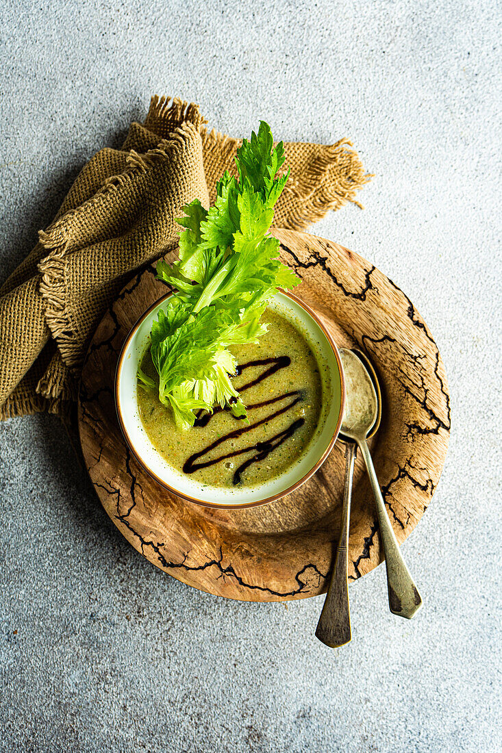 Top view of healthy celery cream soup in bowl with celery sticks served on wooden plate with vintage spoons on grey table next to napkin