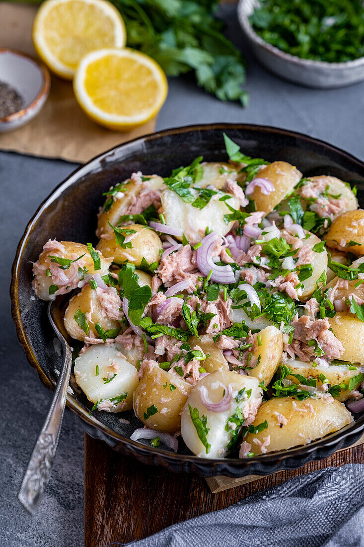 Potato salad with tuna, onions and parsley in a black bowl, with a fork inside, lemon halves and parsley behind it