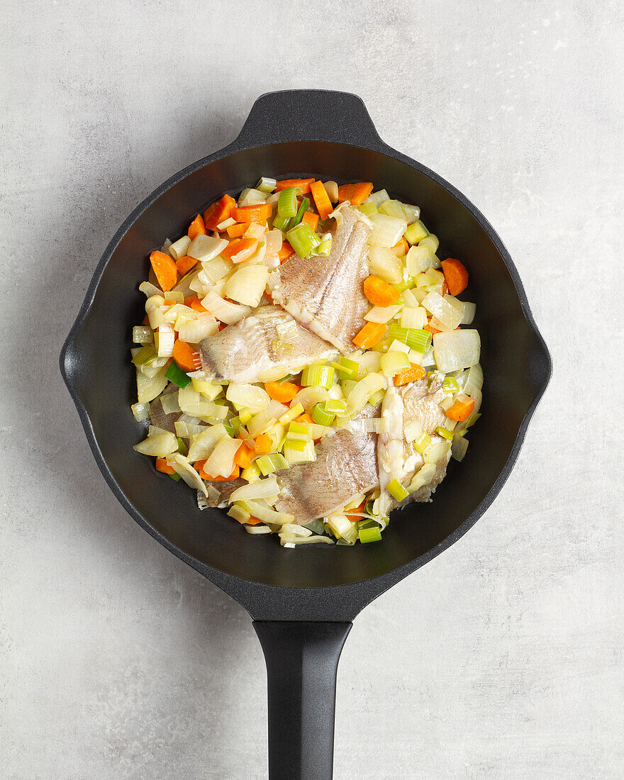 Top view of a frying pan with fumet containing raw, sliced fish and various sliced carrot and cabbage vegetables on a grey table background