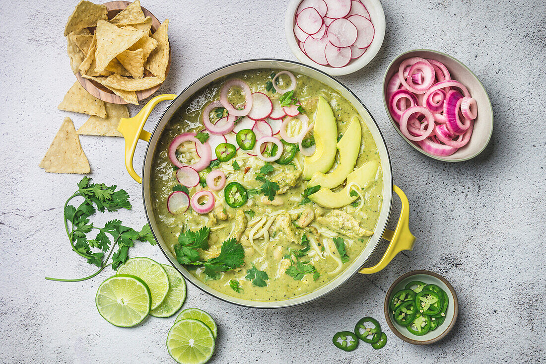 Green chicken chili in yellow pot surrounded by garnishes of chips, jalapenos, picked red onions and radishes