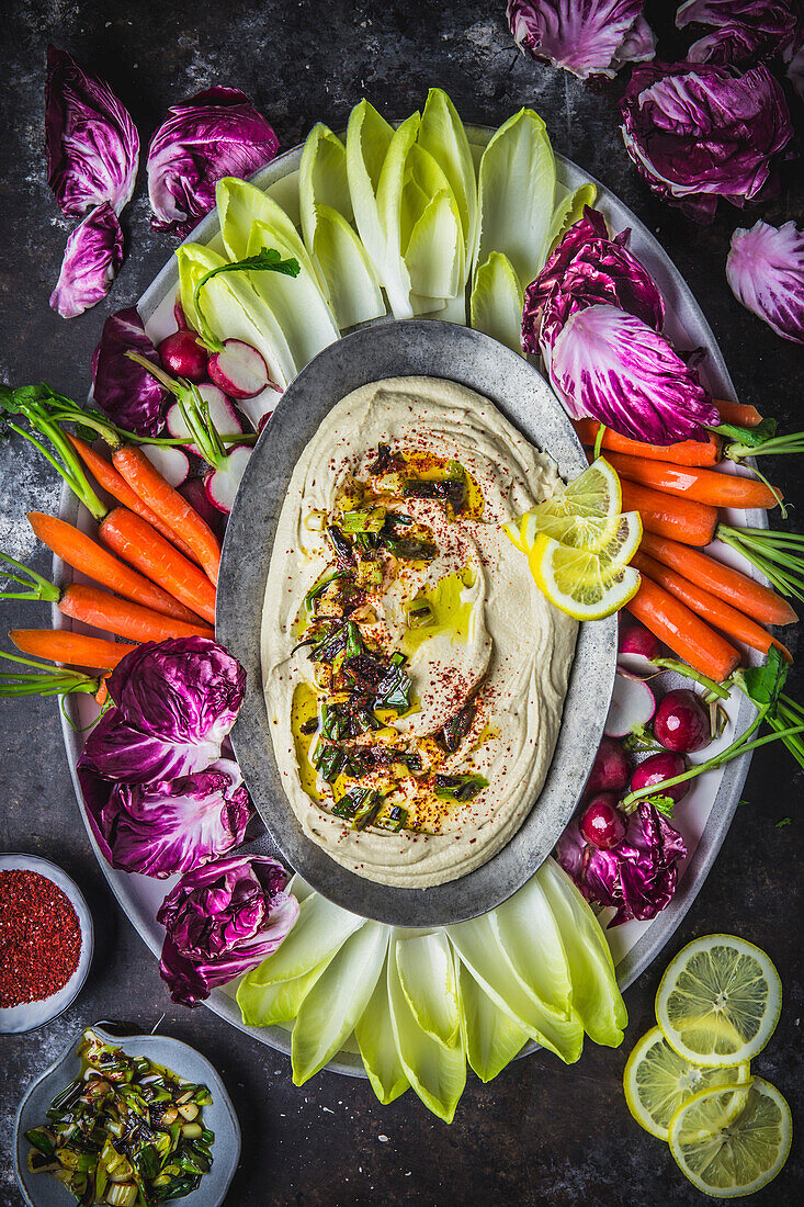 Cremiger Hummus auf ovalem Teller mit Beilagen auf Platte mit buntem Gemüse-Rohkost auf dunklem Hintergrund