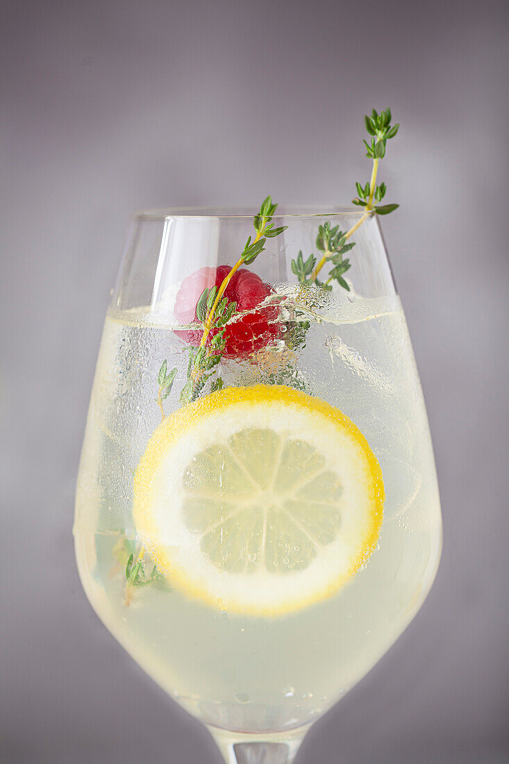 Close-up of a wine glass with a lemon spritzer cocktail in it, garnished with a slice of lemon, raspberry and fresh thyme