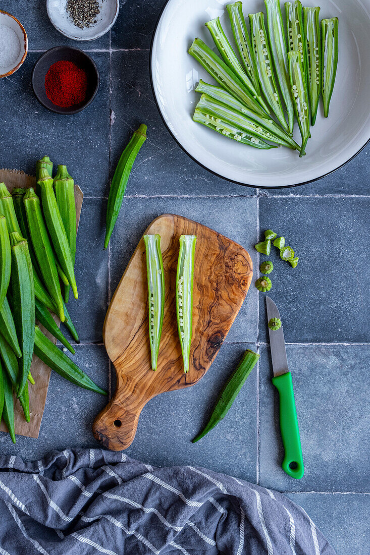 Okra slices in the shape of French fries in a white bowl and an okra pod cut in half lengthways on a chopping board, a knife at the side