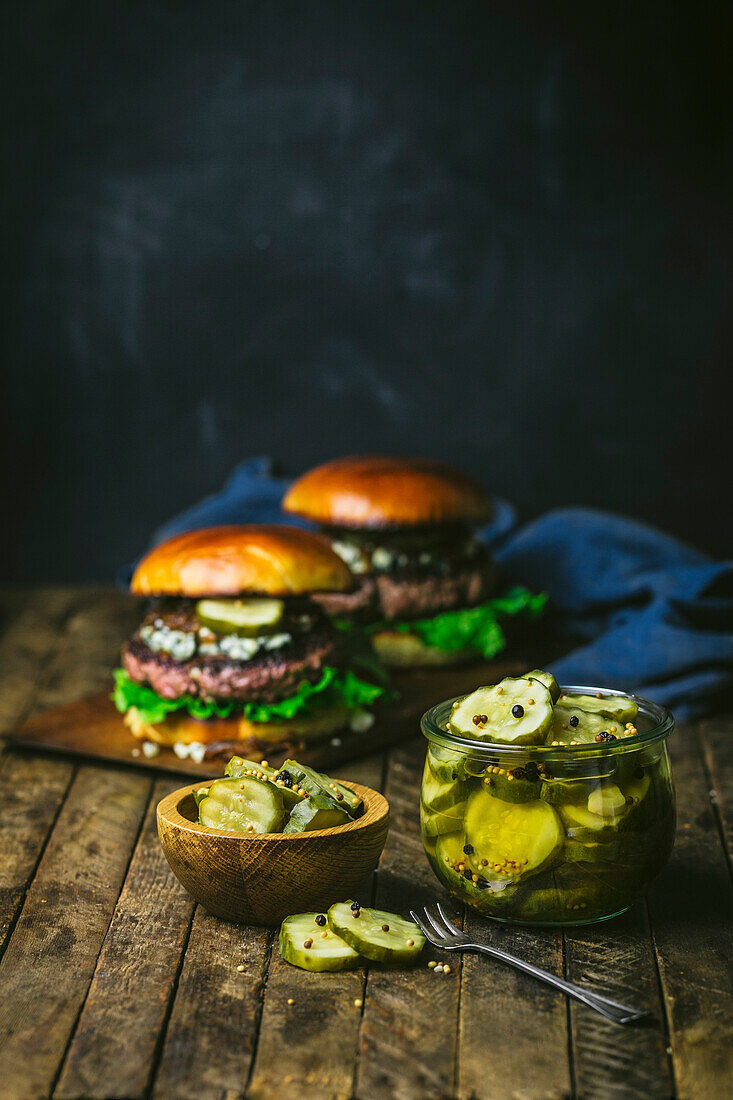 Glass jar of homemade pickle slices, in wood bowl with 2 burgers in background. Wood table and Chalk background
