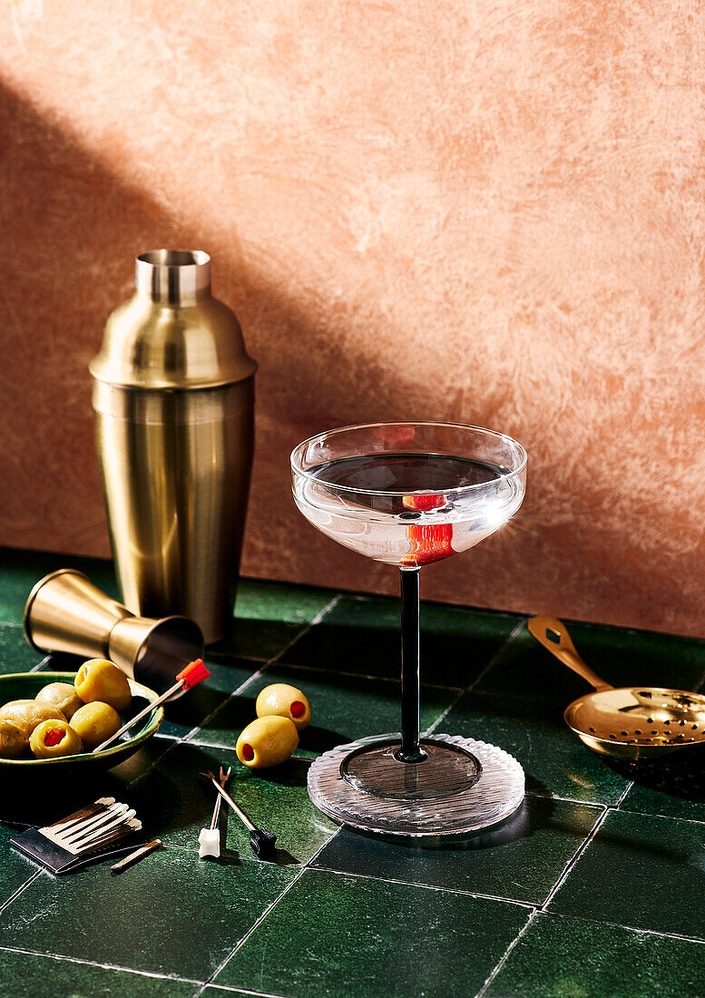 A cocktail glass and a cocktail shaker on a bar with olives