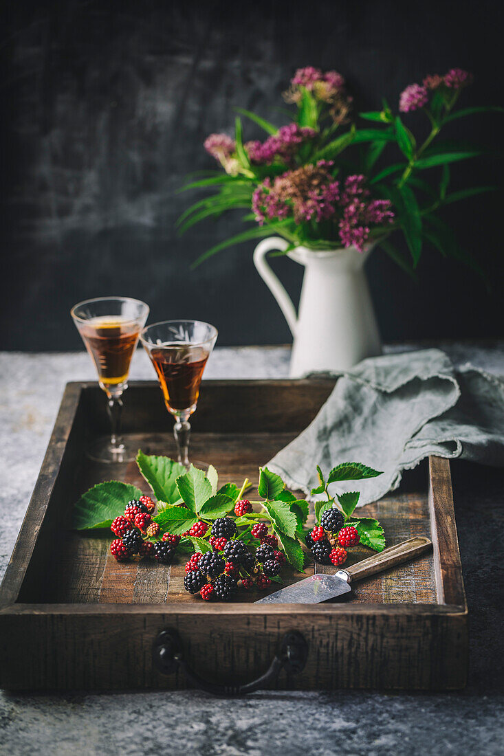 Blackberries and leaves on wooden tray with sherry in glasses and flowers in vase