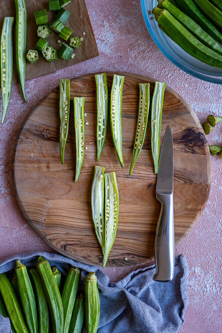 Okra pods are sliced lengthways on a wooden board, a knife lies next to them