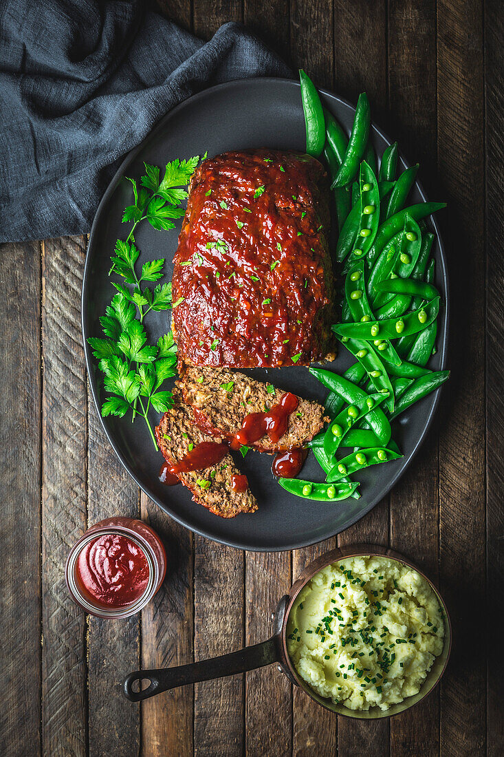 Meatloaf and schnitzel on a platter with fresh sugar snap peas, mashed potatoes in a pan and gravy in a jar