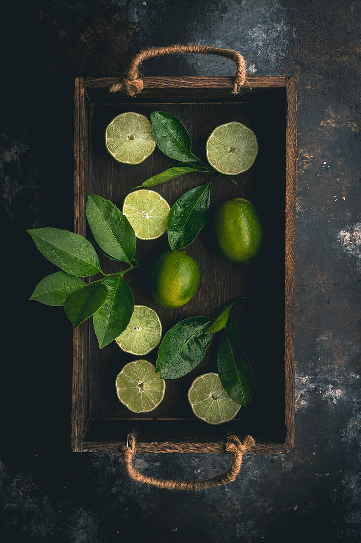 Limes, sliced and whole, and lime leaves in wooden box on dark background