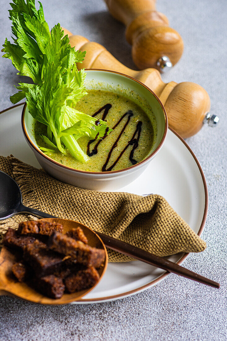 High angle of celery cream soup in bowl with celery sticks on plate with spoon, napkin and bowl bread on table with jars against blurred background