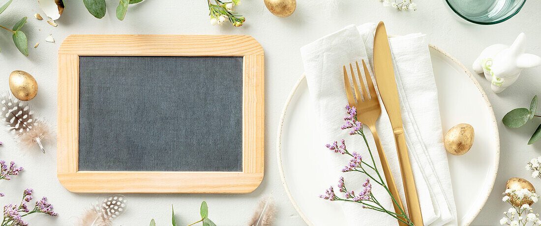 Easter table decorations. Stylish Easter brunch table setting with chalkboard, white plate, napkin, golden cutlery, easter eggs and spring branches on light grey background top view flat lay copy space