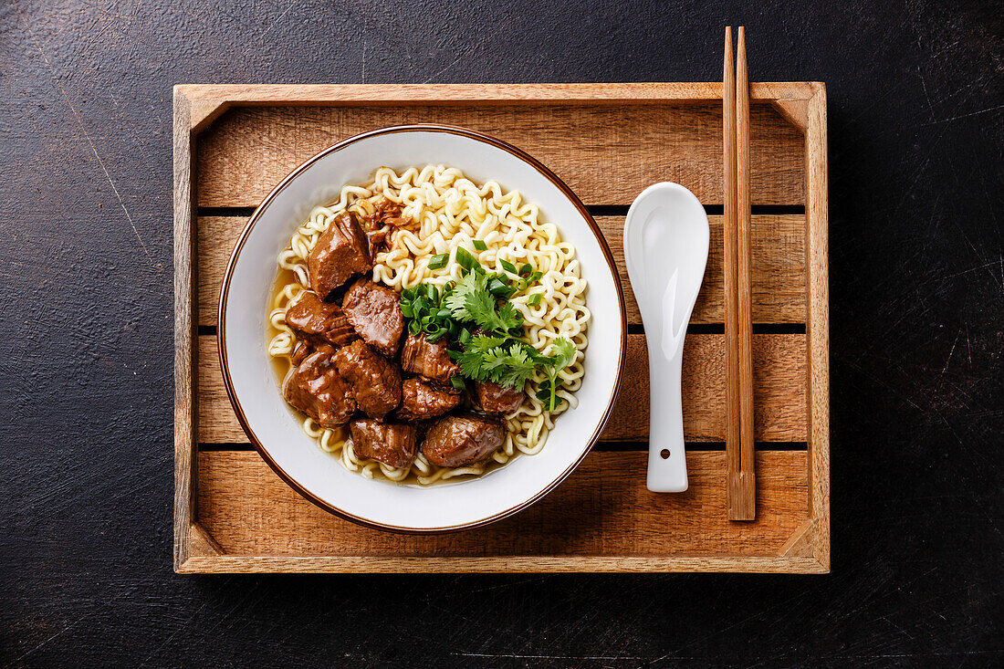 Slow-cooked beef with Asian noodles in broth in wooden tray on a dark background