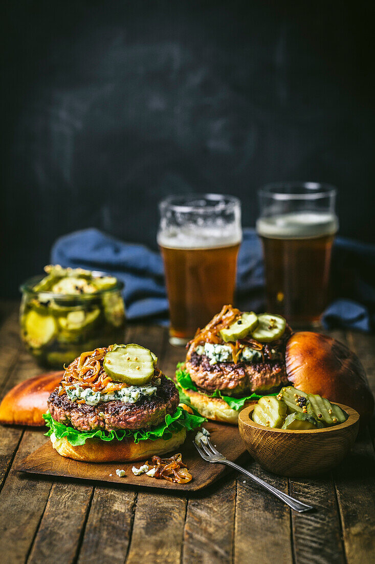 Two beef burgers with sauteed onions, pickles and blue cheese on a wood board with beer pints and pickle jar