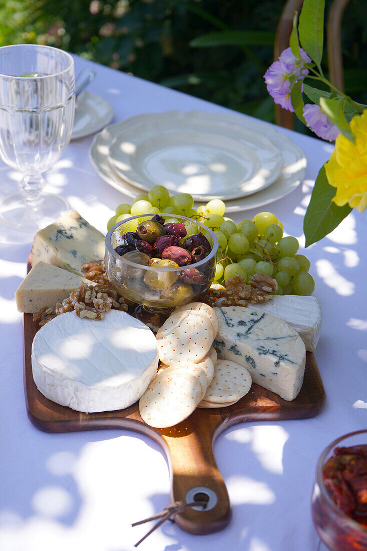 Ein sommerlicher Esstisch im Freien unter einem hohen Baum im gedämpften Schatten. Nahaufnahme einer Käse- und Olivenplatte