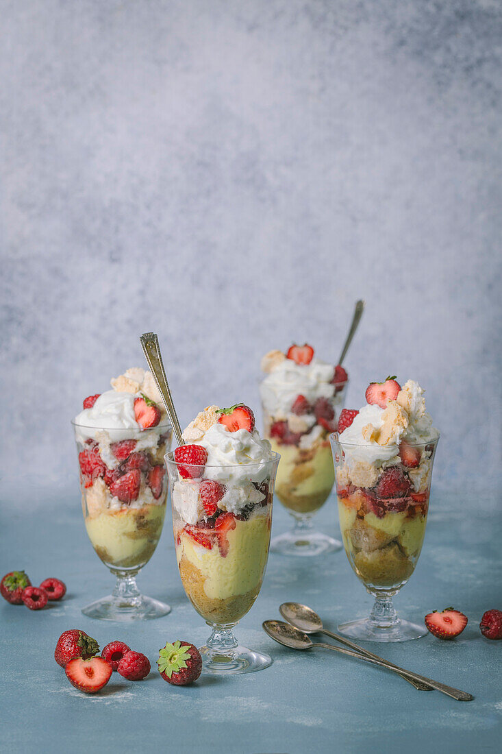 Vier große Parfaitgläser mit Vanillepudding, Kekskrümeln, Schlagsahne und Erdbeeren auf hellblauem Hintergrund