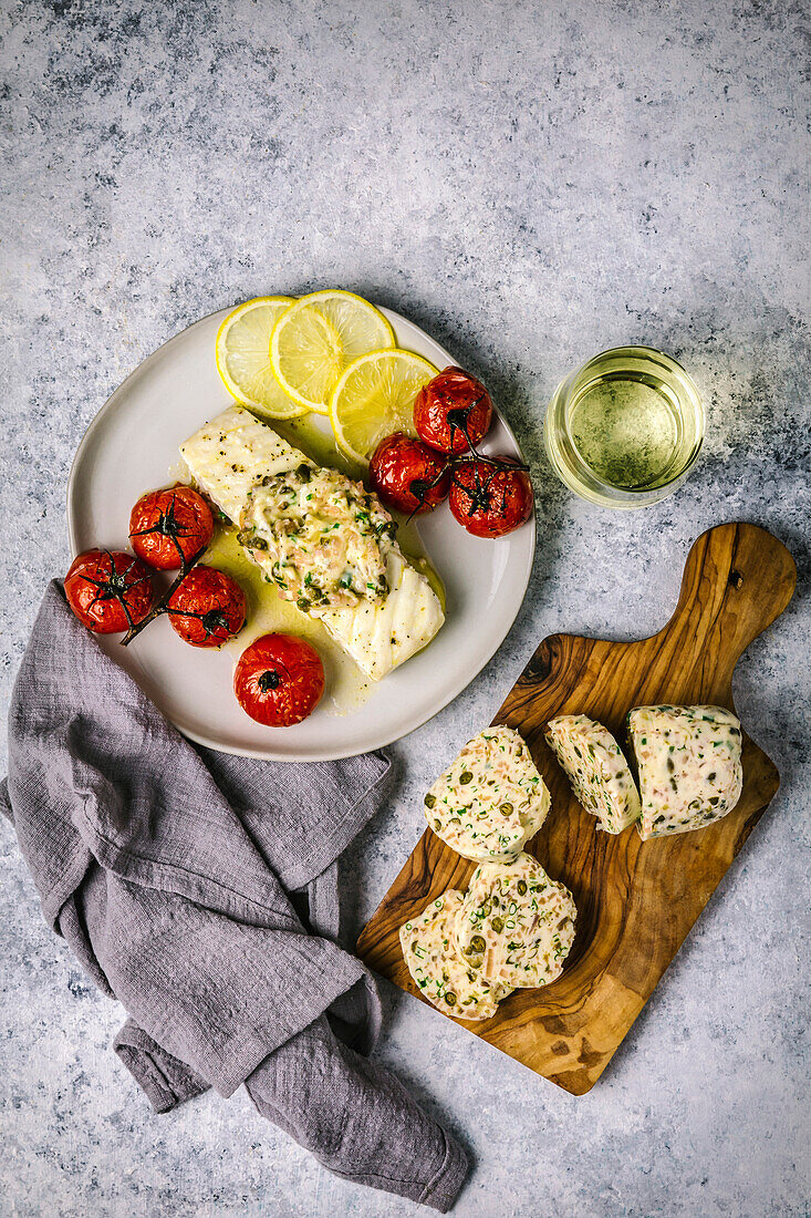 View from above of white fish fillet with melting butter on a white plate with roasted tomatoes, lemon slices and white wine in a stemless glass