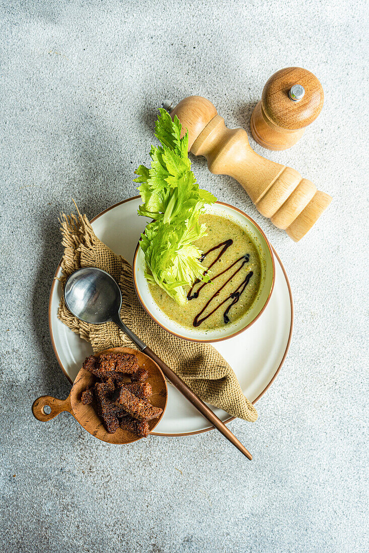Cream of celery soup in a bowl with celery stalks on a plate with a spoon, napkin and bowl with bread on a table with wooden salt and pepper jars, seen from above