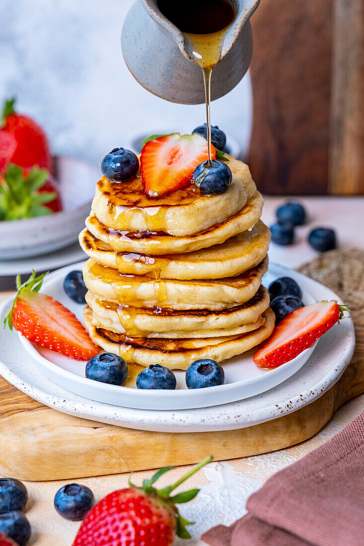 Maple syrup is poured over a stack of pancakes, garnished with blueberries and strawberries