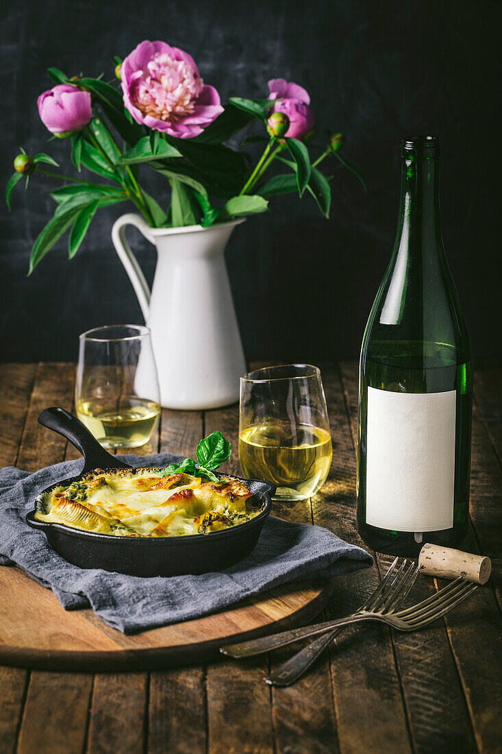 Table setting with baked noodles in a small cast-iron pan with wine glasses, bottles and flowers