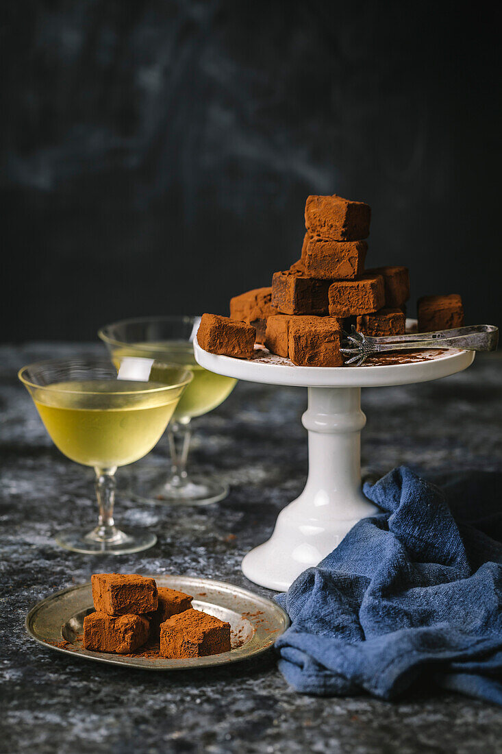Arrangement of chocolate truffles dusted with cocoa powder on a cake stand with a set of silver tongs, and small silver plate with 2 vintage glasses filled with chilled dessert wine