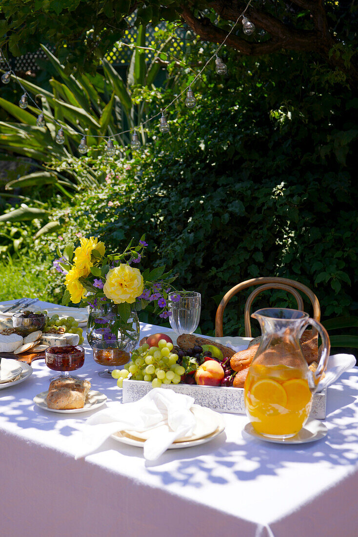 Al fresco summer outdoor dining table laid with fruit, bread tray and cheese platter, in dappled shade under a tall tree.